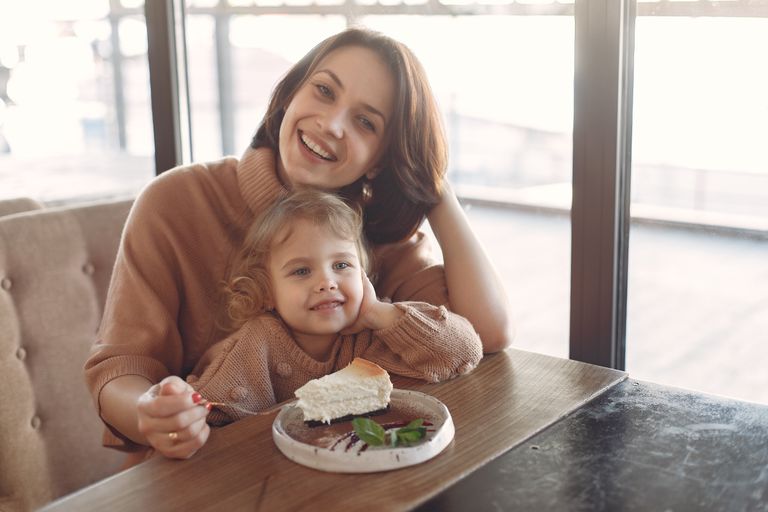 Young woman in casual outfit sitting at desk and eating piece of cake together with little adorable girl while spending time at cafeteria and smiling looking at camera